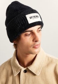Svart stickad mössa med en texturerad yta, prydd med en rektangulär vit lapp märkt "OFF THE WALL", bärs med en beige kordjakett.