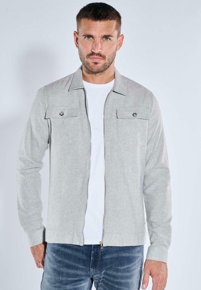 Graue Reißverschlussjacke aus weichem Material, mit zwei Klappentaschen und Kragen, kombiniert mit einem weißen T-Shirt und blauen Jeans.