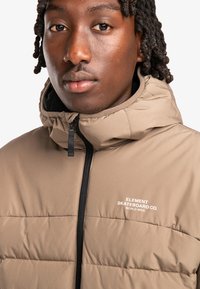 Brown puffer jacket with a high collar, front zipper, and black lining. Features white logo text on the chest and a quilted design.