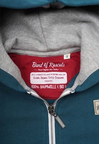 Zip-up hoodie med en teal ytterdel och grå insida, tillverkad av 100% ekologisk bomull, med en röd etikett som anger skötselråd.