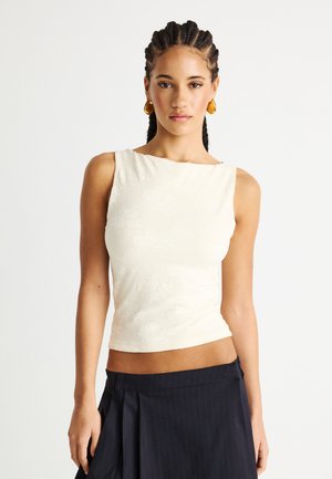 Γυναίκα με πλεξούδες, που φορά χρυσά σκουλαρίκια, αμάνικο λευκό τοπ με σχέδια και σκούρα πλισέ φούστα, στέκεται μπροστά σε απλό φόντο.