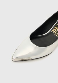 Ασημένια γόβα γυναικεία με μυτερή μύτη, μαύρο εσωτερικό και λογότυπο "Versace Jeans Couture" στη σόλα και μεταλλική άκρη.