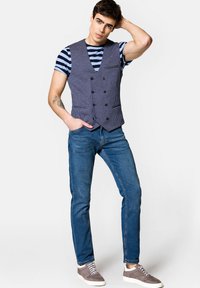 Kamizelka w serek w granatową kratę houndstooth, podwójnie zapinana na czarne guziki, noszona na pasiastym T-shircie; zestawiona z niebieskimi dżinsami i szarymi butami.