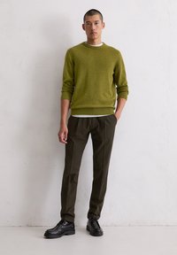 Grüner Wollpullover mit gerippten Bündchen, kombiniert mit dunkelbraunen, maßgeschneiderten Hosen und schwarzen Schnürschuhen vor einem hellen Hintergrund.