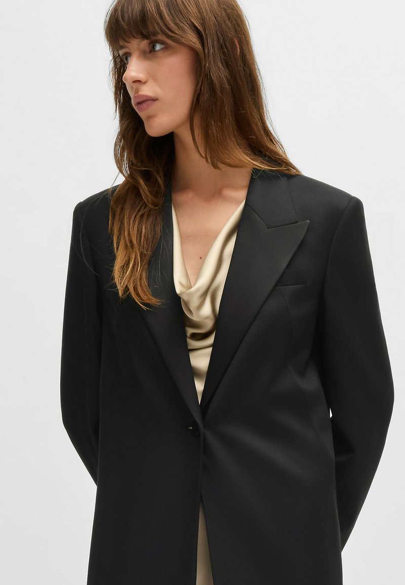 Blazer noir cintré avec un design structuré, des revers nets et une texture lisse. Porté par-dessus un haut en soie léger et drapé.