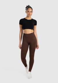 Top corto nero e leggings marroni a vita alta. Il materiale sembra elastico con una texture liscia. Modella che indossa sneakers bianche.