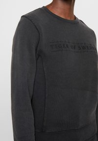 Felpa girocollo grigio scuro con testo ricamato "TIGER OF SWEDEN" sul petto, caratterizzata da un pannello laterale testurizzato e polsini a costine.