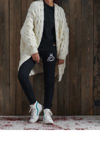 Cárdigan de punto grueso en crema, top ajustado negro, pantalones de chándal negros con logo, zapatillas blancas con acentos turquesa, de pie sobre un suelo texturizado.