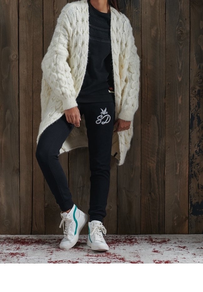 Cárdigan de punto grueso en crema, top ajustado negro, pantalones de chándal negros con logo, zapatillas blancas con acentos turquesa, de pie sobre un suelo texturizado.