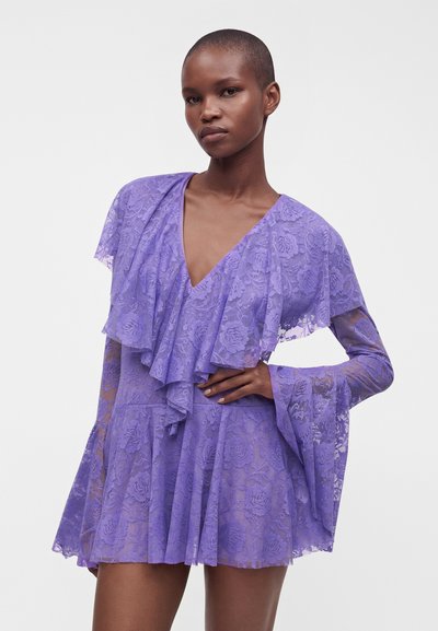 Femme portant une robe en dentelle violette avec de longues manches cloche, un col en V à volants et un motif floral sur un fond clair uni.