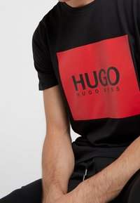 Černé bavlněné tričko s velkým červeným obdélníkovým grafickým prvkem s nápisem "HUGO" v černém písmu. Krátké rukávy a kulatý výstřih.