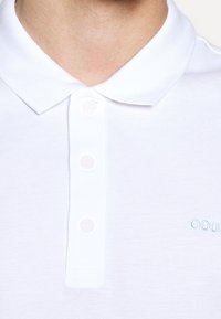 Polo blanc avec une patte de boutonnage à deux boutons et un col plié. Logo brodé discret en bleu clair sur la poitrine gauche. Texture lisse.