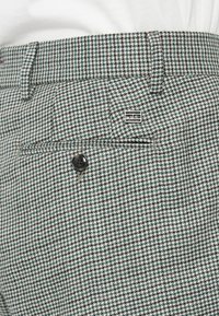 Zöld, fekete és fehér hounds-tooth mintájú szabott nadrág, gombos hátsó zseb, övharapások és egy kis téglalap alakú fém logó kitűző.