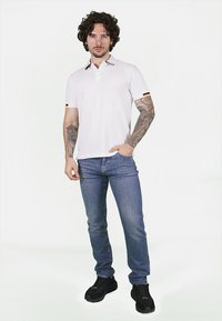 Polo bianco con colletto che presenta accenti gialli e neri, abbinato a jeans blu e sneaker nere. Tatuaggi visibili sulle braccia.