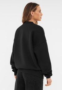 Svart oversized sweatshirt med rund halsringning, långa ärmar, ribbade muddar och en mjuk textur. Sedd bakifrån, mot en vit bakgrund.
