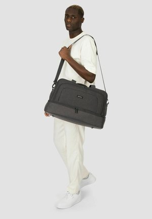 Hombre con atuendo blanco llevando un gran bolso de hombro gris oscuro con la etiqueta "REDOLZ" sobre un fondo liso.