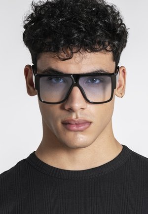 Mladý muž s kučeravými vlasmi, ktorý nosí veľké štvorcové čierne slnečné okuliare a malý kruhový náušník, oblečený v čiernom rebrovanom tričku.