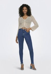 Top corto beige lavorato a maglia con nodo frontale, motivo strutturato e maniche lunghe, abbinato a jeans aderenti blu e sandali con tacco azzurri.