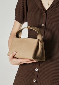 Bež torba od kože sa strukturiranim oblikom, kratkom ručkom i suptilnom teksturom. Dizajn uključuje elegantan, minimalizam stil.