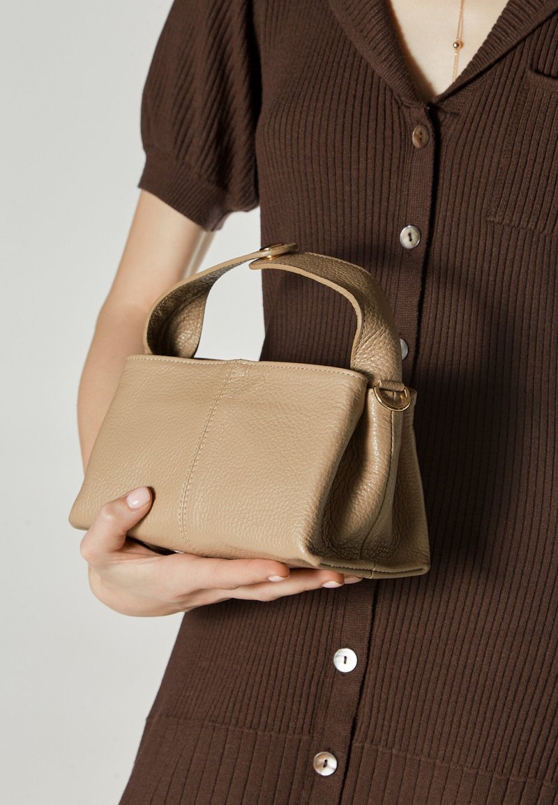 Bež torba od kože sa strukturiranim oblikom, kratkom ručkom i suptilnom teksturom. Dizajn uključuje elegantan, minimalizam stil.