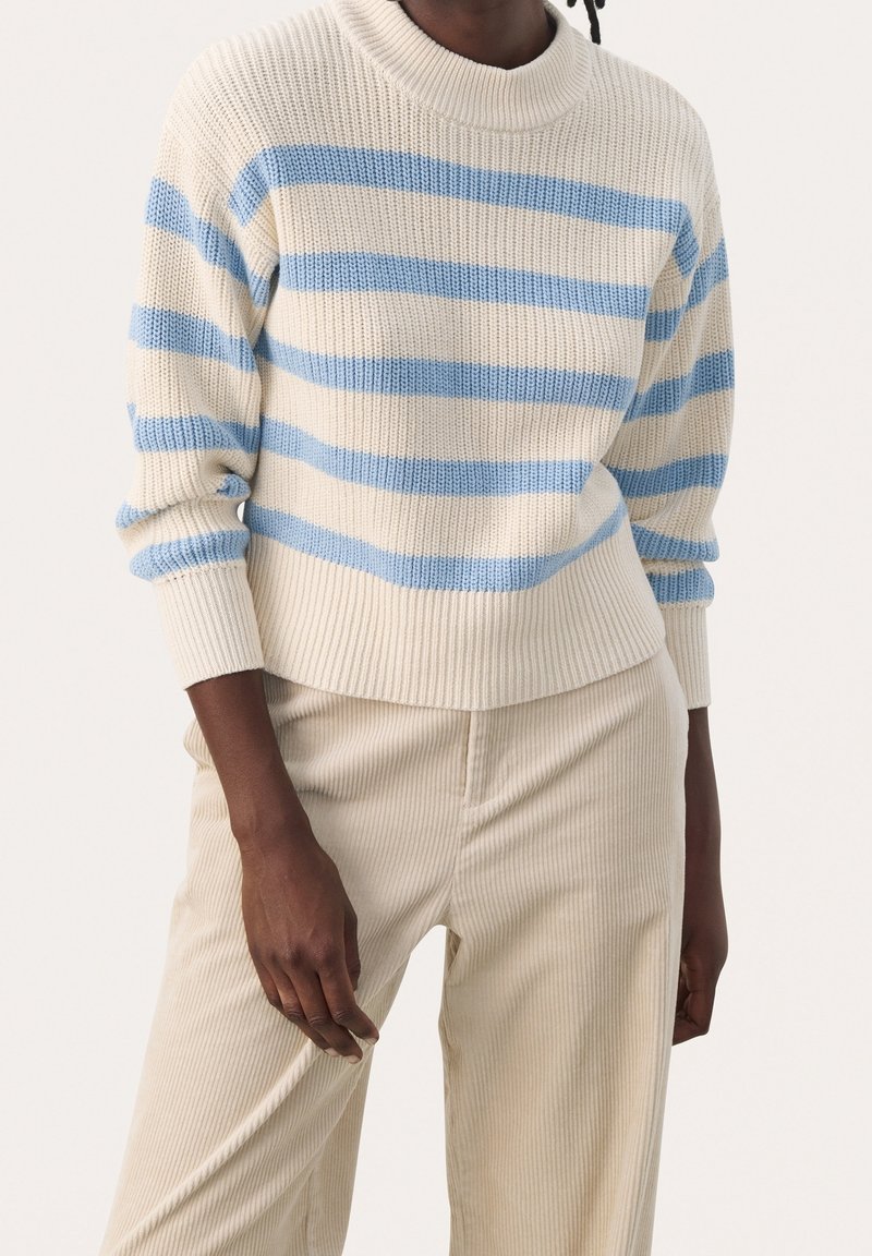 Cremefarbener Strickpullover mit blauen Horizontalstreifen, mit einem gerippten Ausschnitt und Bündchen. Kombiniert mit hellbeigen Cordhosen.