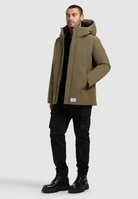 Olive green insulated jacket with a hood, front zipper, side pockets, worn over a black shirt and paired with black cargo pants and boots.