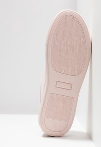 Semelle d'une basket rose clair avec un motif de sculptures ondulées, reposant verticalement contre un coin mural et sol blanc.