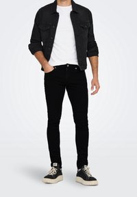 Chaqueta de mezclilla negra con dos bolsillos frontales, llevada sobre una camiseta blanca. Combinada con jeans negros ajustados y zapatillas negras con suelas blancas.
