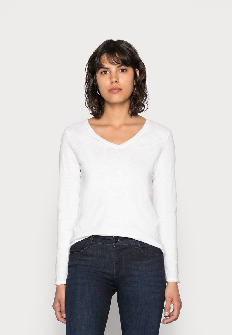 Camisa blanca de manga larga con escote en V, fabricada en una tela suave, presenta un corte relajado y una sutil textura, combinada con jeans de mezclilla oscura.