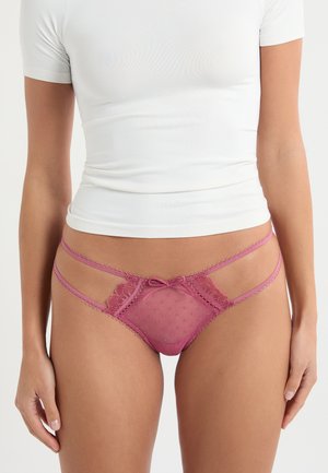 Donna che indossa un perizoma in pizzo rosa trasparente con doppie fascette laterali e un piccolo fiocco al centro, abbinato a un top bianco aderente e corto.