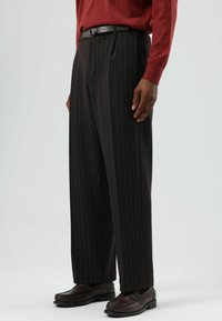 Zwarte broek met krijtstreep en plooien, gemaakt van een glad materiaal. Gecombineerd met een rood shirt met lange mouwen en bruine loafers.