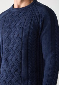 Dunkelblauer Strickpullover mit einem strukturierten Zopfmuster und geripptem Ausschnitt; hat glatte Seitenpaneele für einen Kontrast.