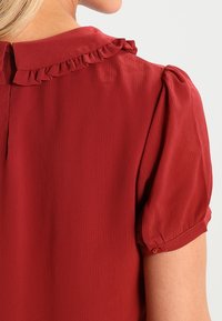 Blouse rouge texturée avec col volanté, manches bouffantes et poignets à boutons. Le tissu présente un motif côtelé subtil.