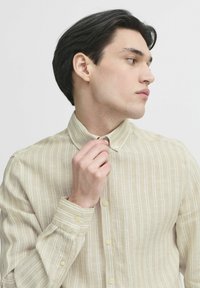 Beige stripete skjorte med lange ermer og krage med knapper, svak tekstur og lyse knapper. Modellen justerer kragen med den ene hånden.