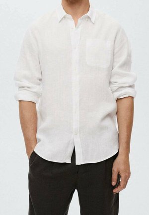 Hombre con una camisa de lino blanca abotonada con las mangas remangadas y pantalones negros, con las manos en los bolsillos, contra un fondo liso.