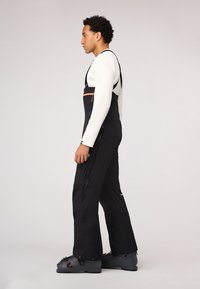 Schwarze Skioveralls mit verstellbaren Trägern, ausgestattet mit einem orangen Reißverschluss-Brusttasche. Strukturierter Stoff und seitliche Reißverschlussöffnungen erhöhen die Funktionalität.