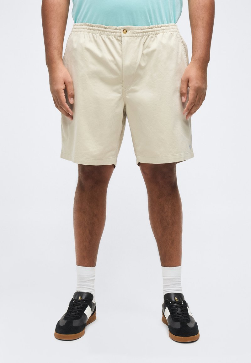 Άτομο που φοράει μπεζ σορτς με ελαστική μέση, ανοιχτό γαλάζιο πουκάμισο, λευκές κάλτσες και μαύρα και λευκά παπούτσια, στέκεται σε απλό φόντο.