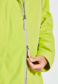 Limegrüne Jacke mit Reißverschluss aus glattem Stoff. Sie verfügt über einen kontrastierenden weißen Reißverschluss und eine Hand, die den Reißverschlussgriff hält.