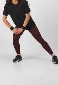 Černé tričko, bordó legíny, černo-růžové sportovní boty, černé zápěstní pásky. Model v protažené pozici na jednobarevném pozadí.