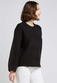 Un pull en laine noir avec un col rond et des manches longues, présentant une coupe décontractée et un ourlet côtelé. Un jean blanc avec des accents floraux jaunes.