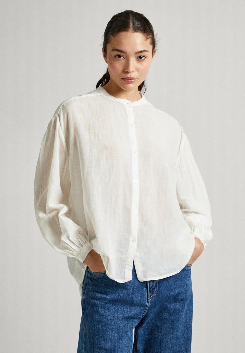 Camicia bianca a maniche lunghe con bottoni, realizzata in tessuto leggero, con colletto rotondo, maniche voluminose e una vestibilità morbida. Abbinata a jeans blu.