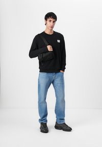 Felpa nera con un piccolo logo, jeans blu e sneaker nere. L'individuo porta una borsa a tracolla nera.