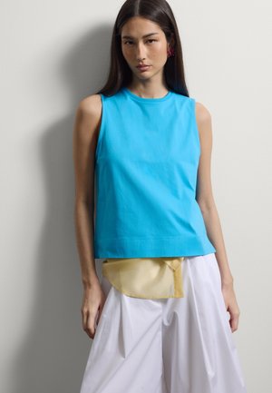 Junge Frau mit langen dunklen Haaren trägt ein ärmelloses, leuchtend blaues Oberteil, lockere weiße Hose und ein gelbes Stoffdetail an der Taille, steht an einer Wand.