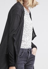 Cardigan leggero nero indossato sopra una camicia bianca con un motivo a triangoli neri sparsi, abbinato a jeans grigio scuro.