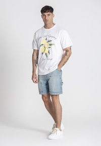 Biela bavlnená tričko s grafikou citróna a textovým dizajnom, spárované s svetlomodrými džínsovými šortkami a bielymi teniskami s béžovými šnúrkami.