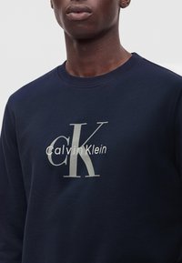 Homme portant un sweat-shirt bleu marine avec de grandes lettres gris clair "cK" et un texte "Calvin Klein" à l'avant.