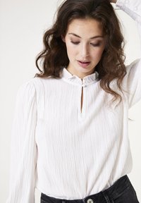 Witte textuurblouse met een gerimpelde kraag en een keyhole-opening, voorzien van lange mouwen en een subtiel gestreept patroon. Model draagt zwarte jeans.