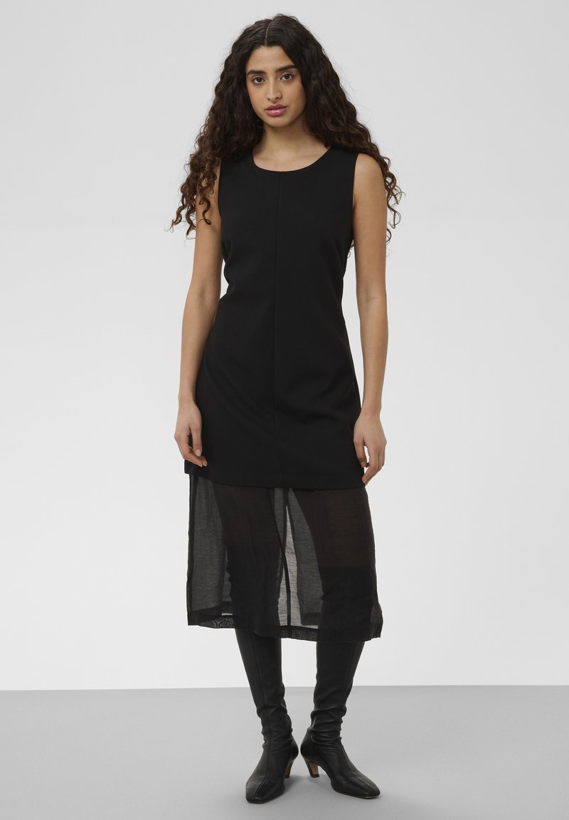Femme aux longs cheveux bouclés portant une robe noire sans manches avec une superposition en tulle sur la jupe et des bottes noires à talons jusqu'aux genoux.