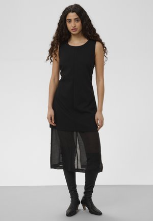 Donna con lunghi capelli ricci che indossa un vestito nero senza maniche con sovrapposizione di gonna trasparente e stivali neri con tacco fino al ginocchio.