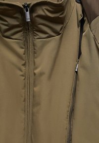 Gros plan sur une veste vert olive avec une fermeture éclair centrale et une poche zippée partiellement ouverte sur le côté droit.